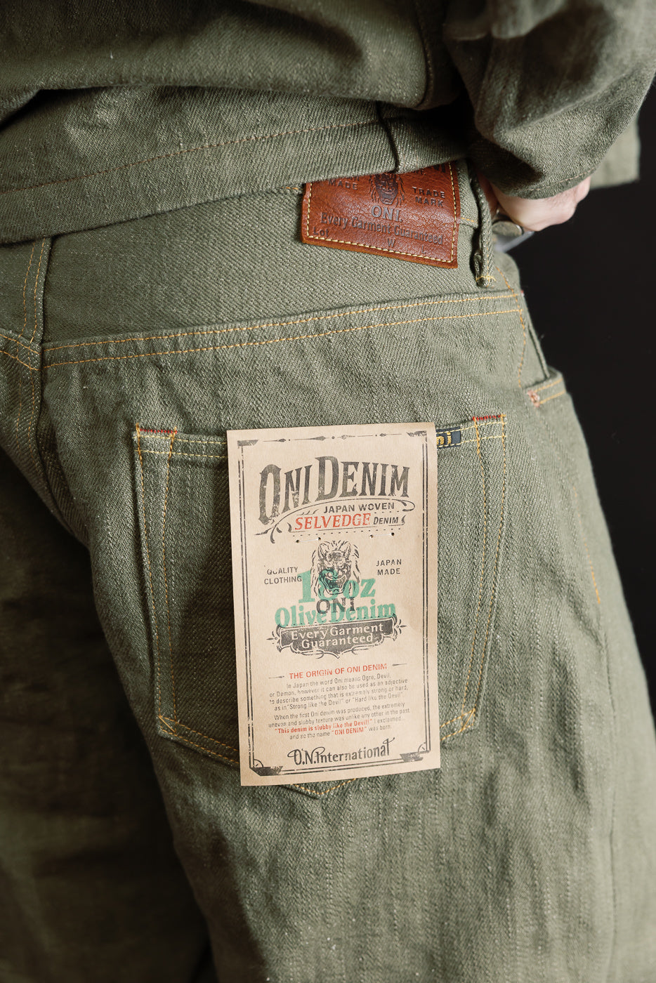 Close-up of male model’s back right side wearing 18oz olive selvedge denim jeans, showing rear pocket, leather patch, and paper tag; model is 5'9