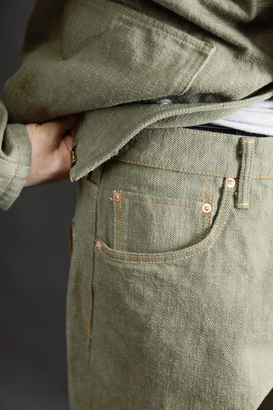 Close-up of front left side of olive selvedge denim jeans on male model, showing coin pocket, rivets, and textured fabric. Model is 5'9