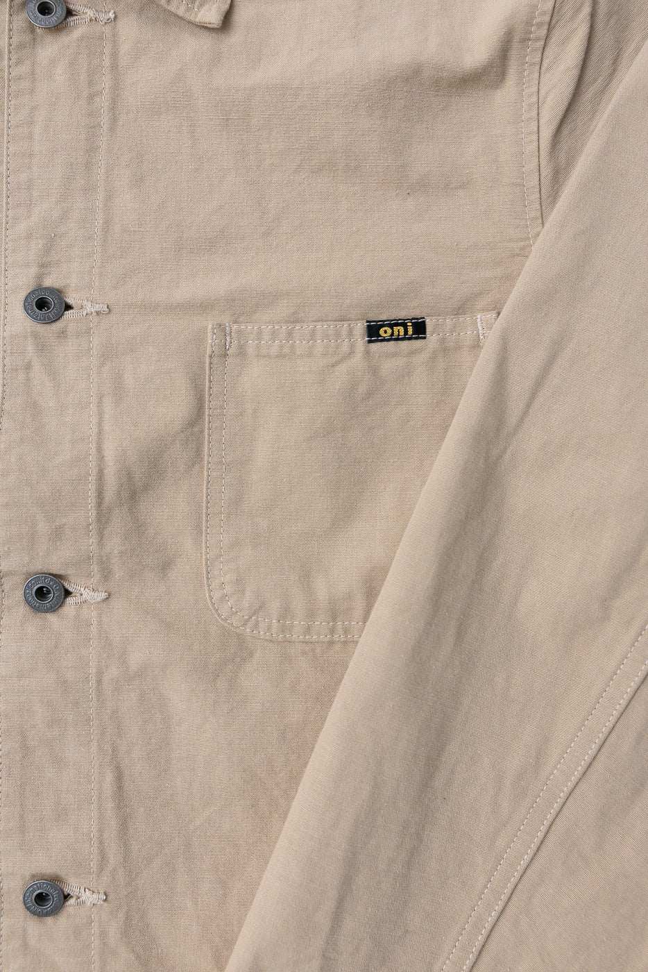 Close-up front view of beige work shirt: chest pocket with 'oni' tag, metal buttons, and visible fabric texture