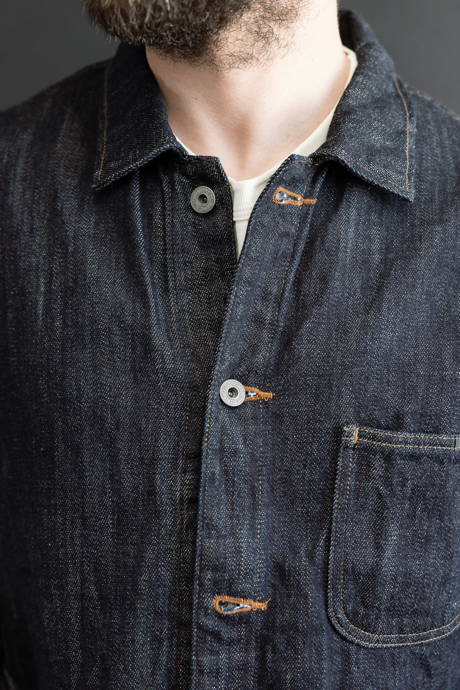 Close-up front view of male model in dark selvedge denim chore coat with button closure, orange stitching, chest pocket