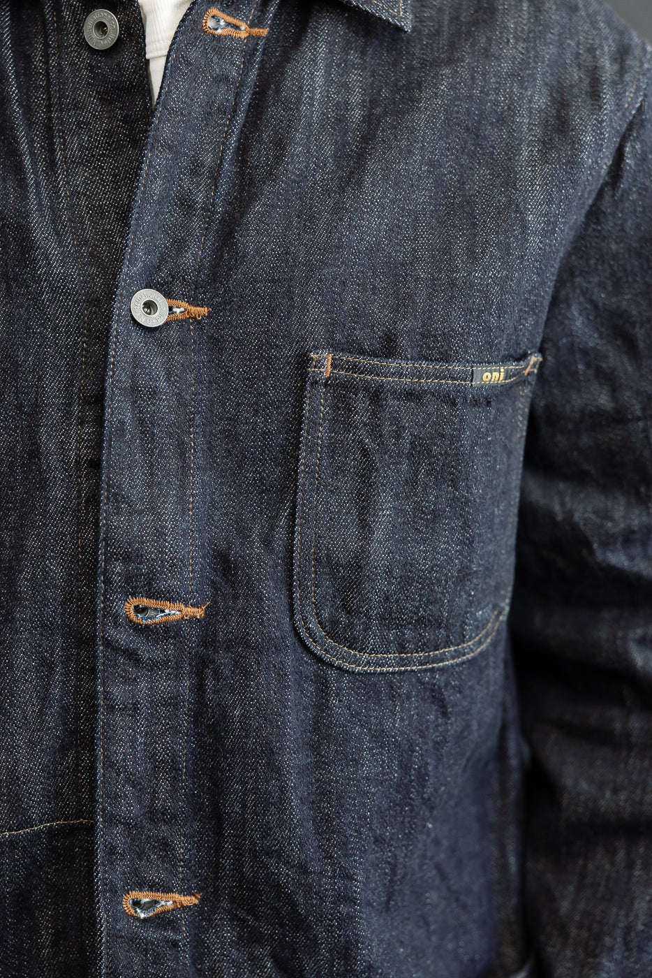 Close-up front view of dark selvedge denim shirt with orange stitching, metal buttons, chest pocket, and tag detail