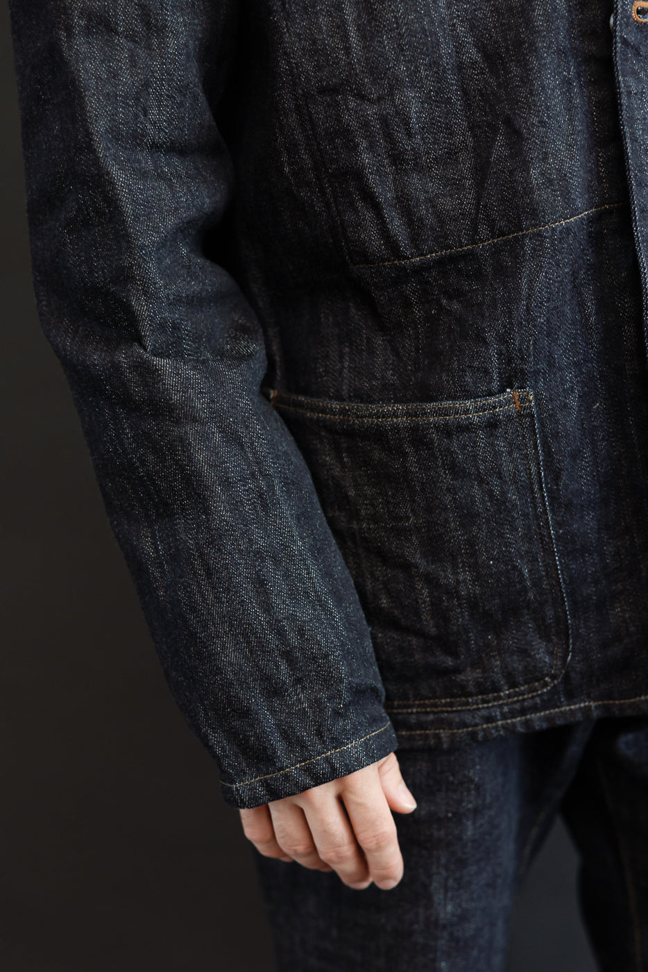 Close-up side view of male model in dark selvedge denim jacket, showing sleeve cuff, pocket detail, and fabric texture
