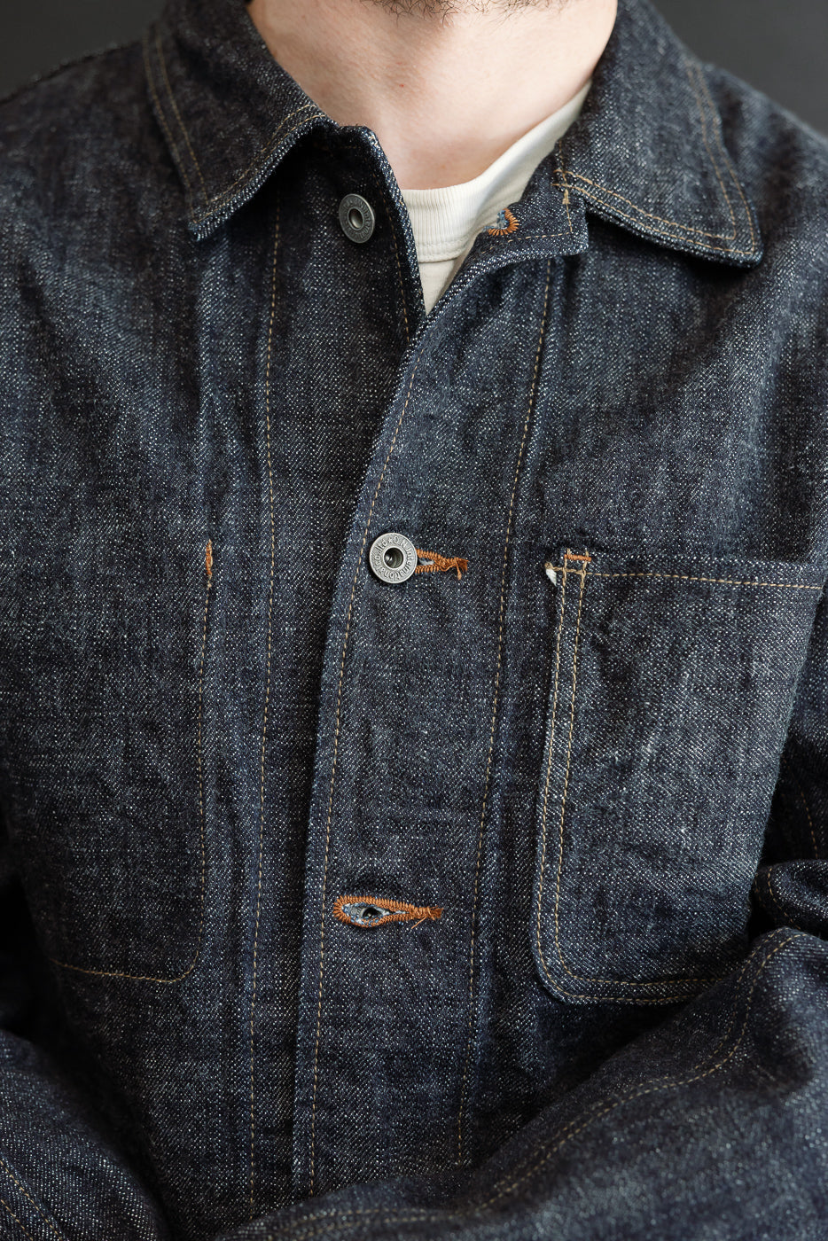 Close-up front view of male model wearing dark denim chore jacket with orange stitching, button closure, and chest pockets.