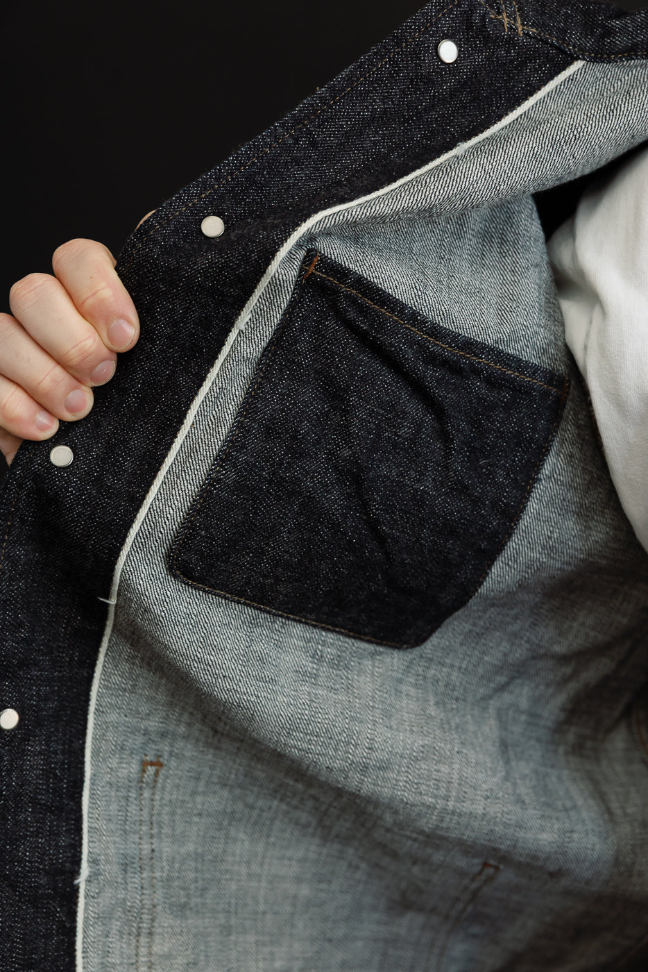 Close-up of dark denim jacket interior, showing chest pocket, white stitching, and button details on male model.