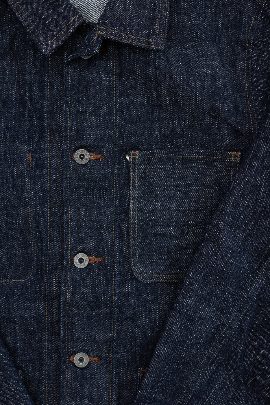 Close-up front view of dark selvedge denim shirt, showing button placket, chest pocket, and orange stitching detail