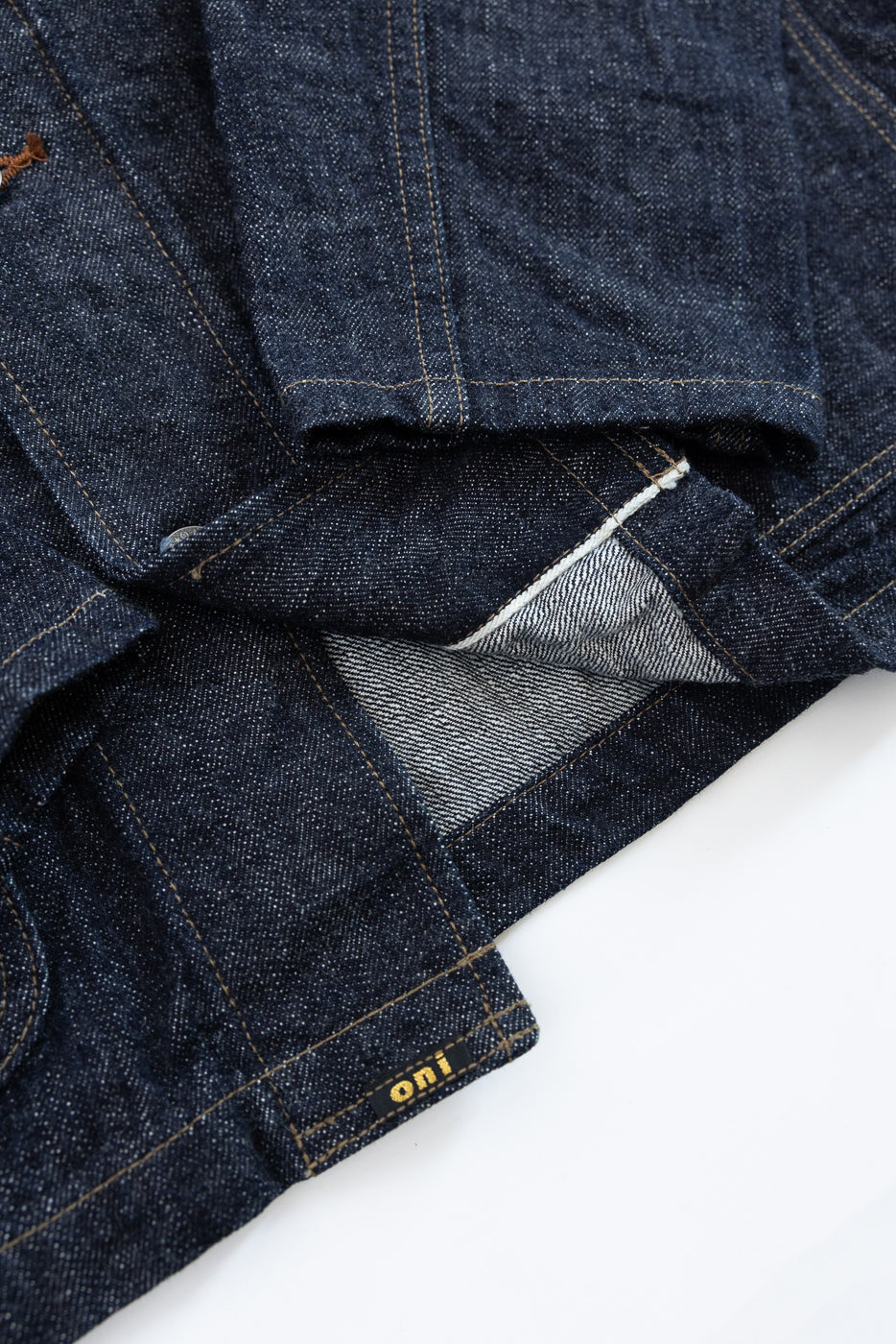 Close-up front view of dark selvedge denim jacket, showing raw edge, contrast stitching, and 'oni' tag detail