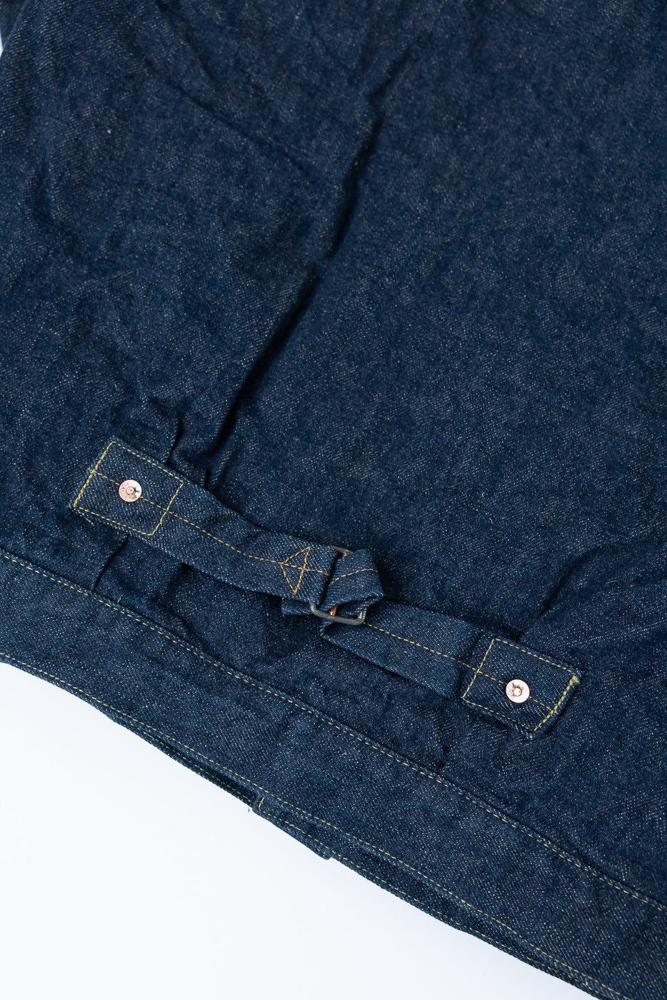 Close-up of back hem and adjustable cinch strap on selvedge denim jacket, showing fabric texture and stitching details.