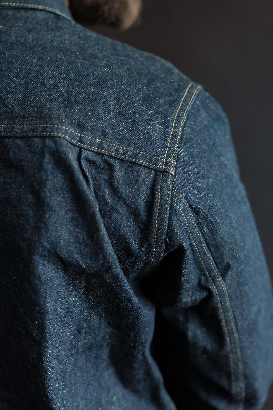 Close-up of back shoulder and sleeve seam on male model wearing selvedge denim jacket, highlighting texture and stitching.