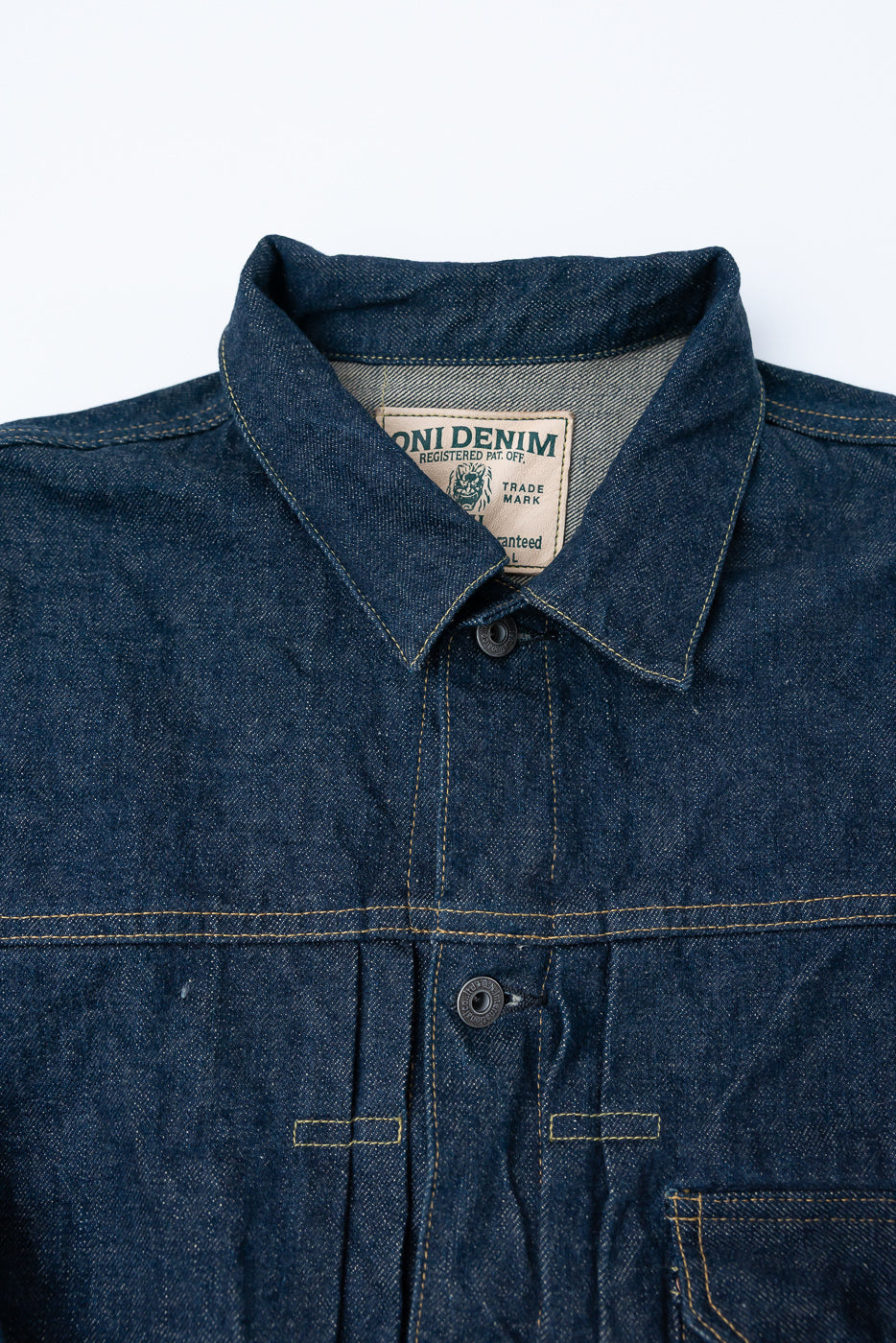 Close-up front view of men's selvedge denim jacket collar, button, chest stitching, and tag details.