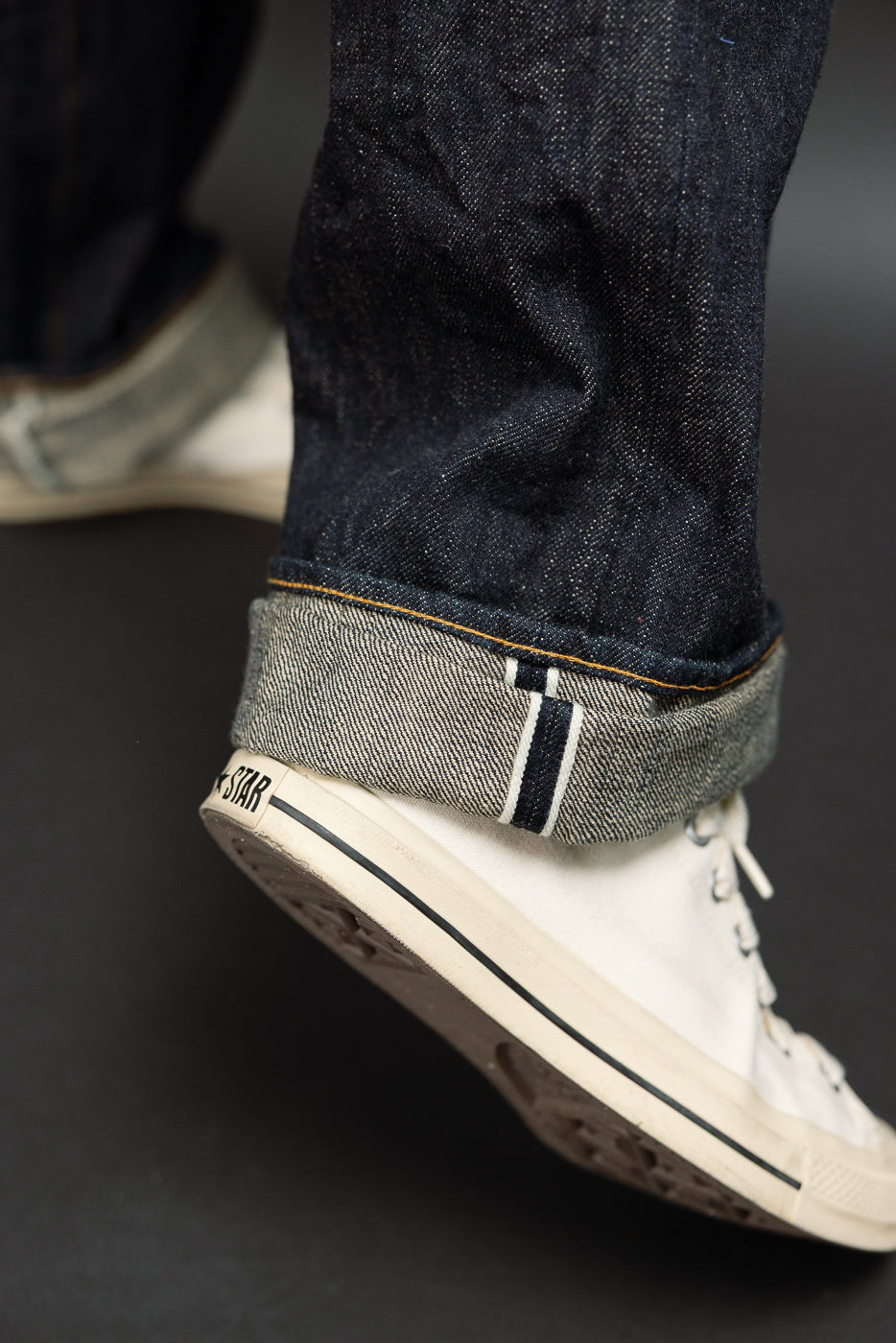 Close-up of male model’s cuffed selvedge denim jeans showing raw hem, contrast stitching, and white sneaker