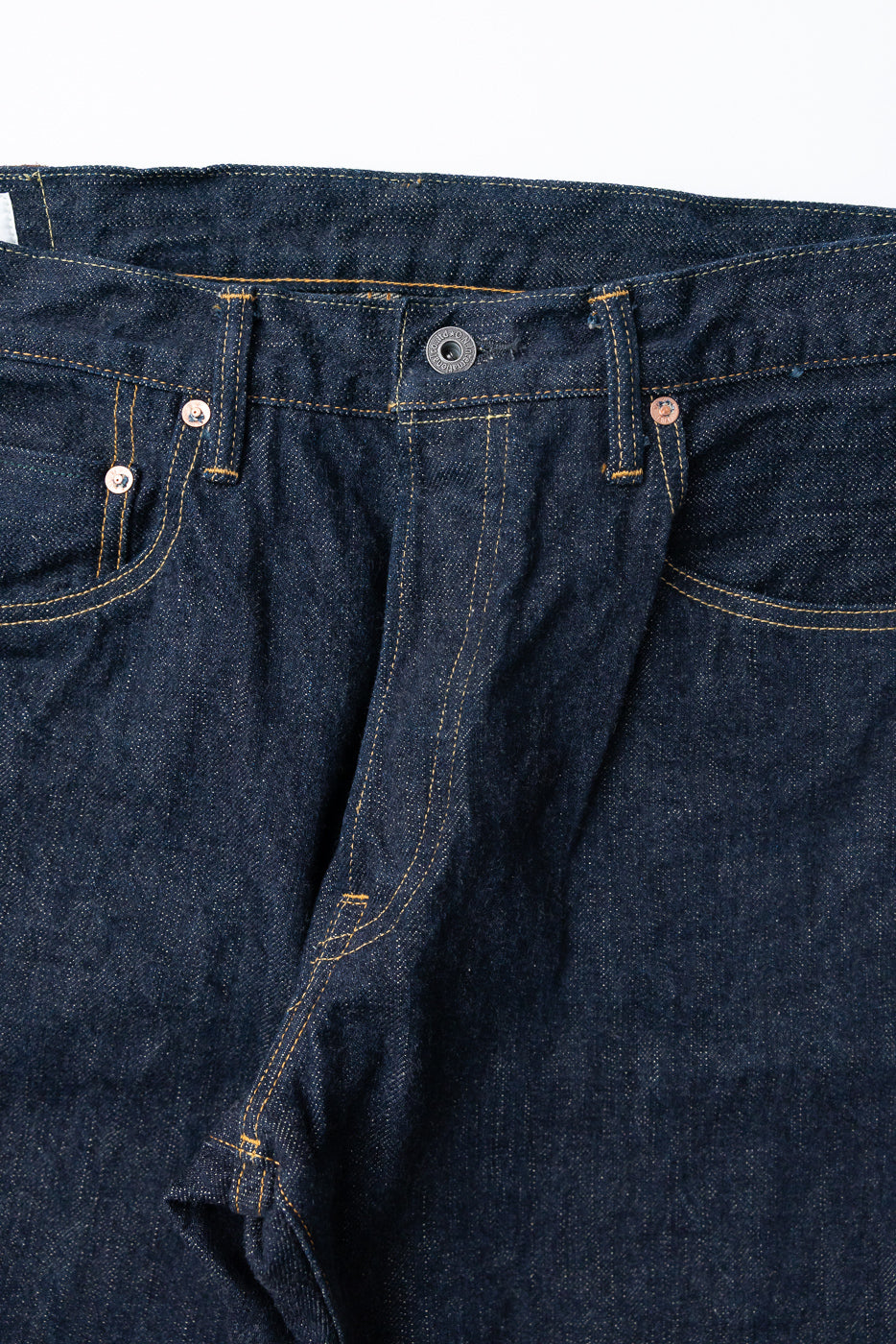 Close-up front view of dark selvedge denim jeans showing button fly, copper rivets, and yellow stitching detail
