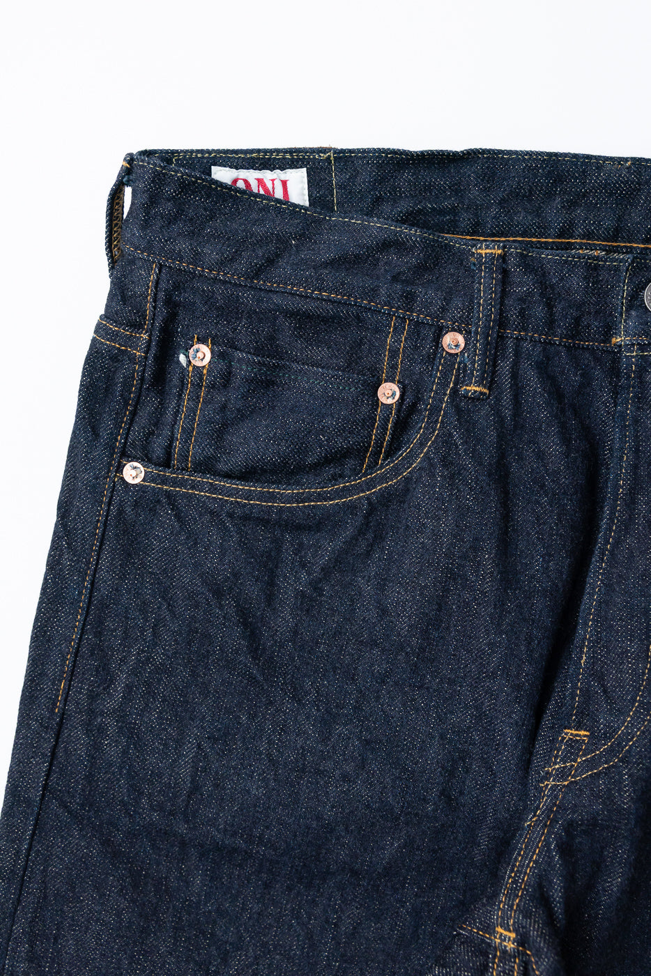 Close-up front view of dark selvedge denim jeans showing back pocket, copper rivets, and waistband tag detail
