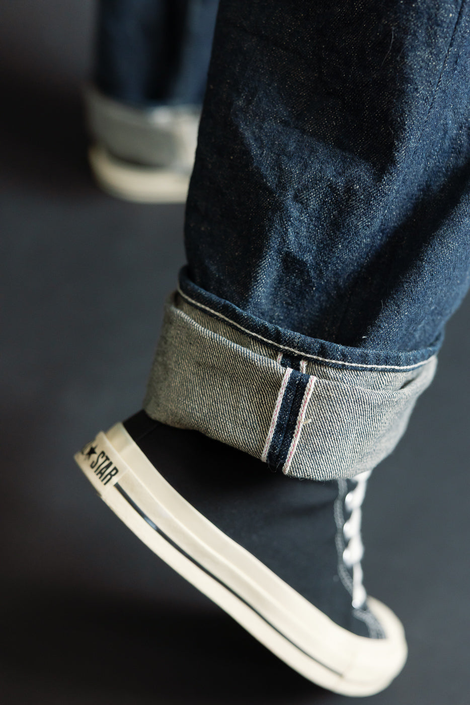 Close-up of male model's cuffed selvedge denim jeans hem, side view, showing fabric texture and selvedge ID. Model is 5'9