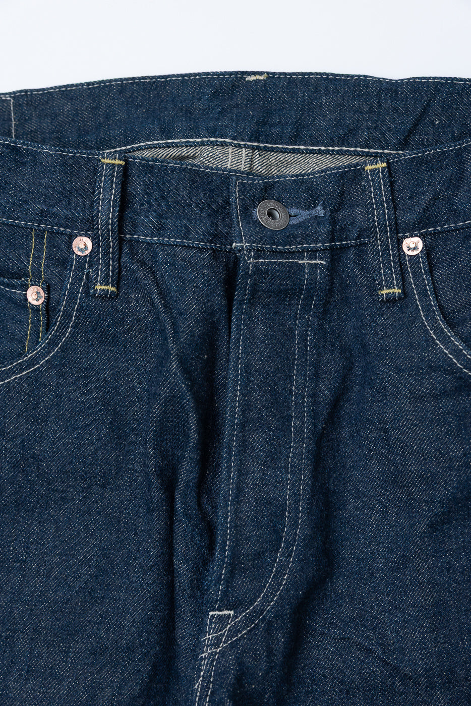 Close-up front view of selvedge denim jeans waist, showing button, rivets, stitching details.