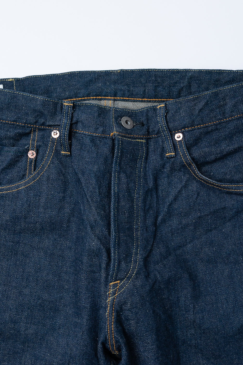 Close-up of front waist and fly of selvedge denim jeans, showing button, rivets, and stitching.
