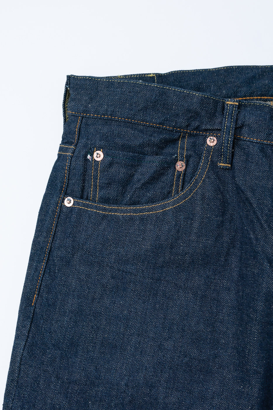 Close-up of front right pocket and waistband on selvedge denim jeans, showing copper rivets and textured indigo fabric.