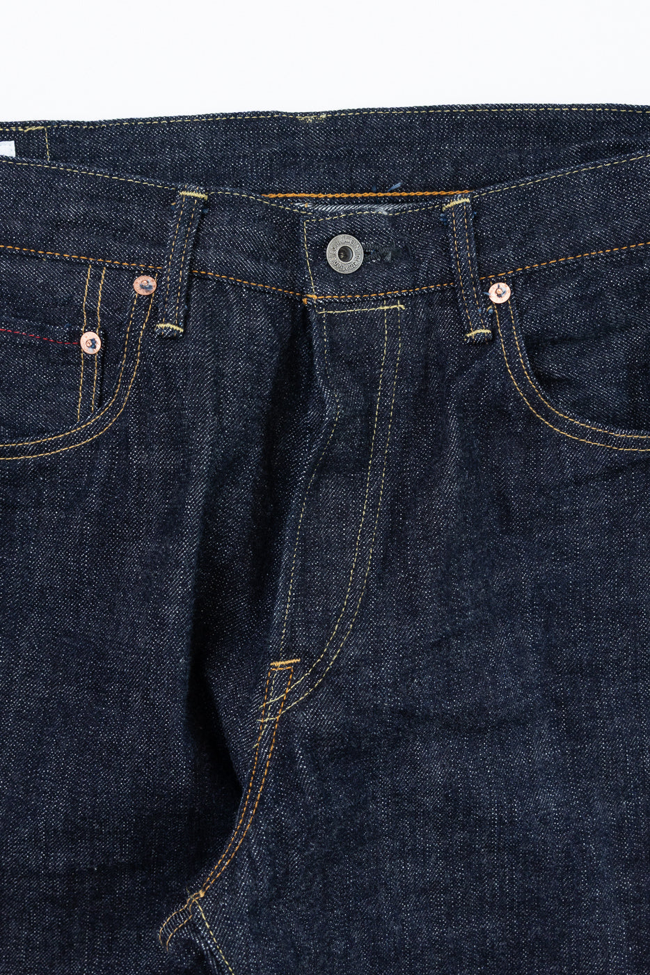 Close-up front view of raw selvedge denim jeans showing copper rivets, contrast stitching, and button fly detail