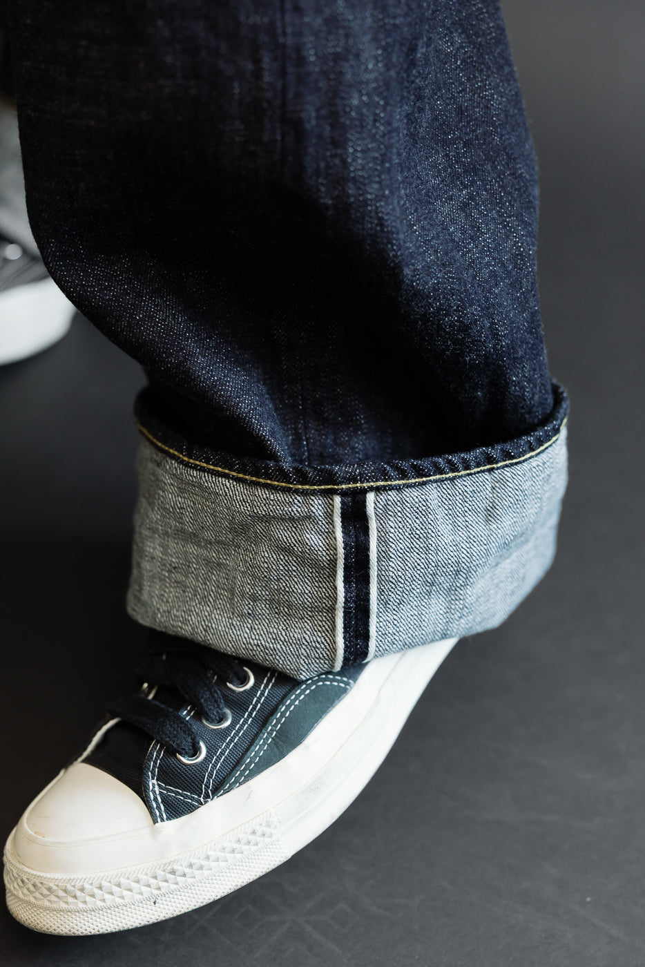 Close-up of male model's cuffed selvedge denim hem, showing raw edge & contrast stitching, paired with dark canvas sneakers