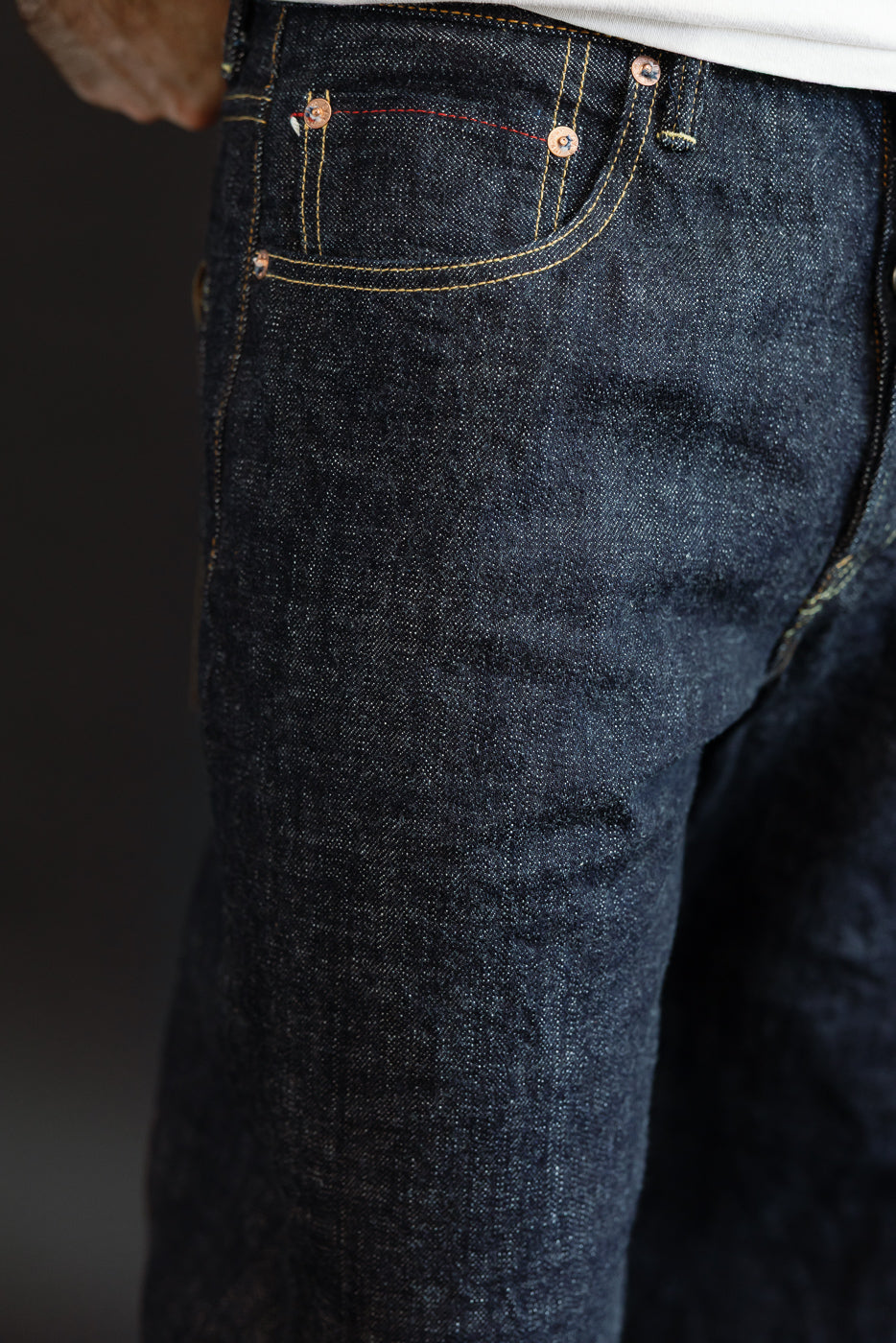 Close-up front view of dark selvedge denim jeans on male model, showing copper rivets, yellow stitching, and fabric texture