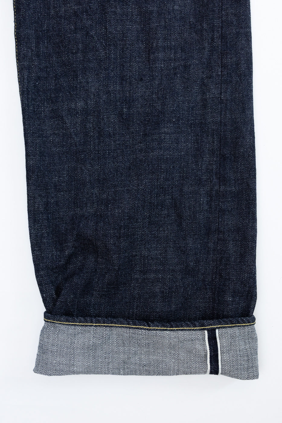 Close-up front view of dark selvedge denim jeans leg, showing raw hem with contrast stitching and selvedge edge detail
