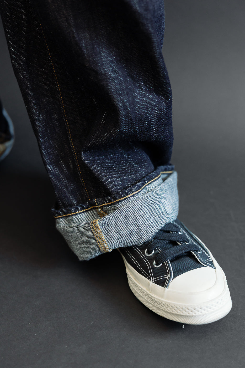 Close-up of male model's cuffed selvedge denim jeans, showing raw hem and contrast stitching with black sneakers