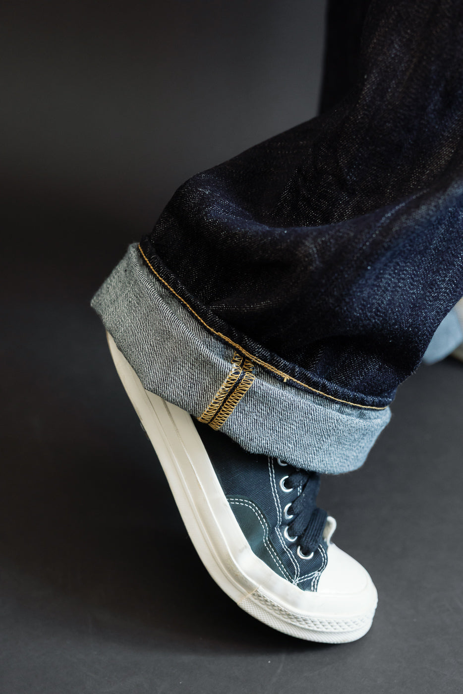 Close-up side view of male model's cuffed selvedge denim jeans showing raw hem, gold stitching, and dark wash with navy sneakers