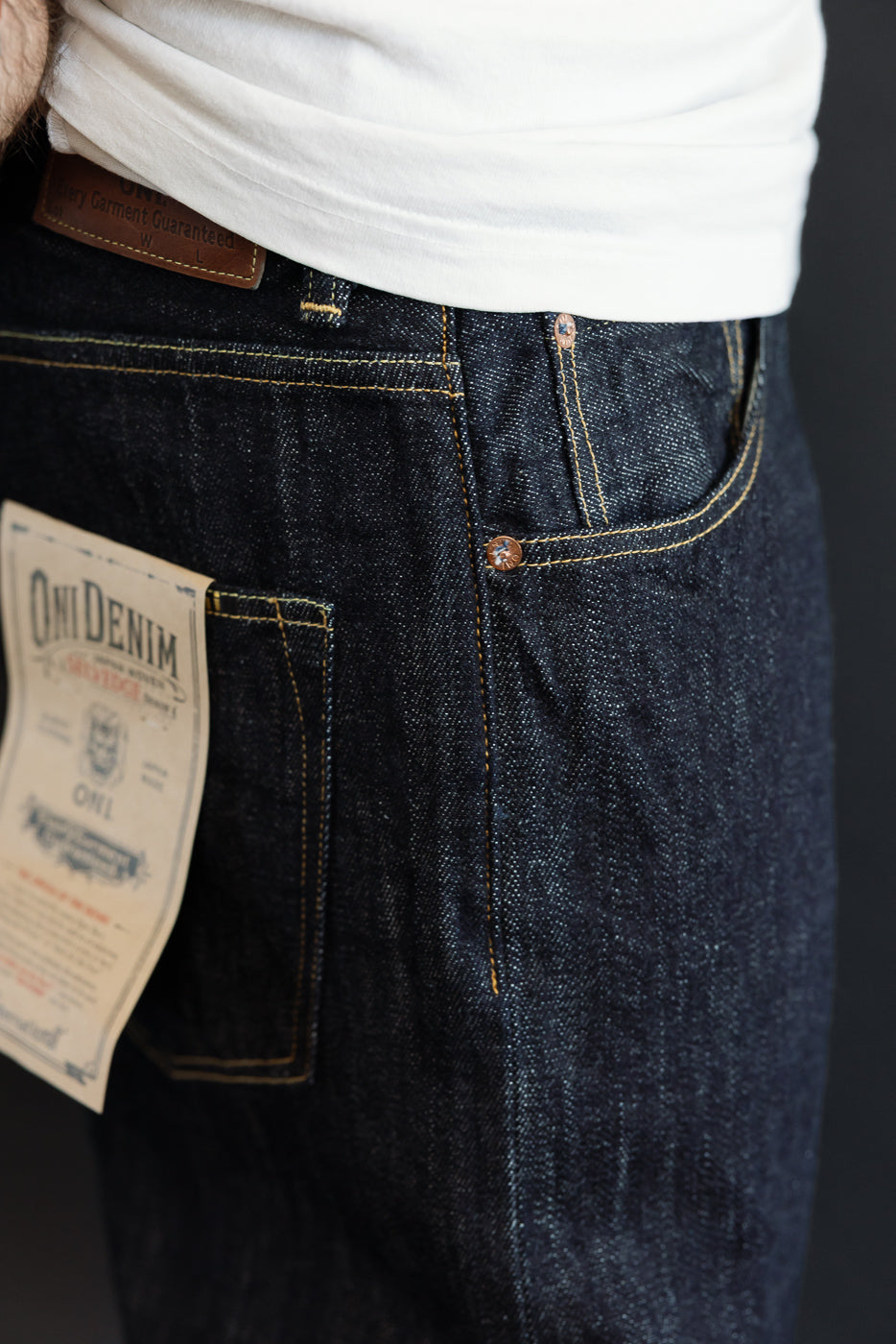 Close-up of male model's back, showcasing dark selvedge denim jeans with gold stitching, branded patch, and copper rivets.