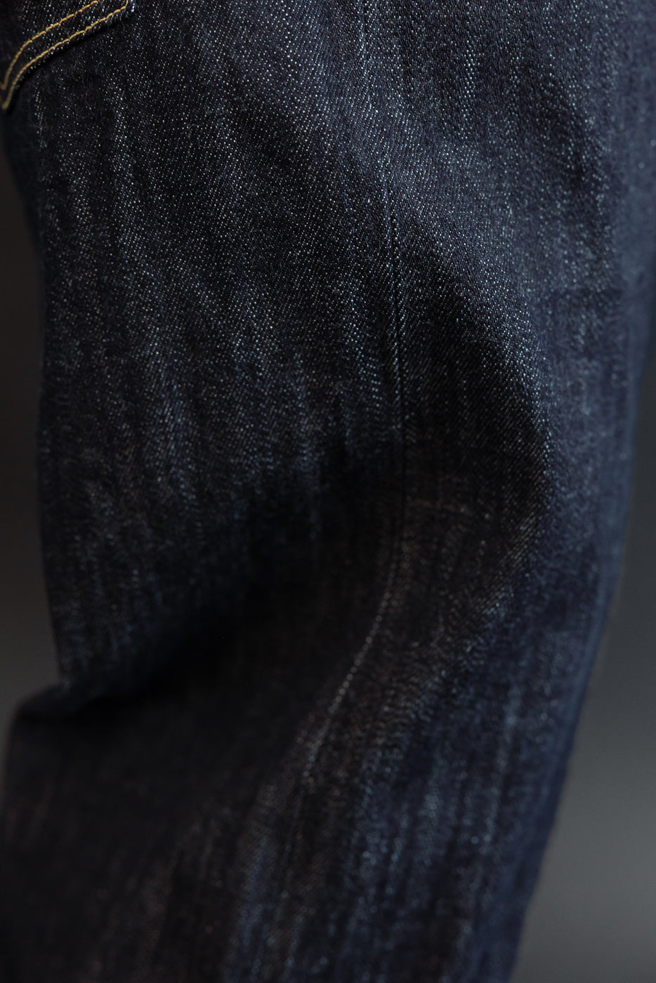 Close-up of dark selvedge denim jeans, front thigh view, showing fabric texture and yellow stitching detail.