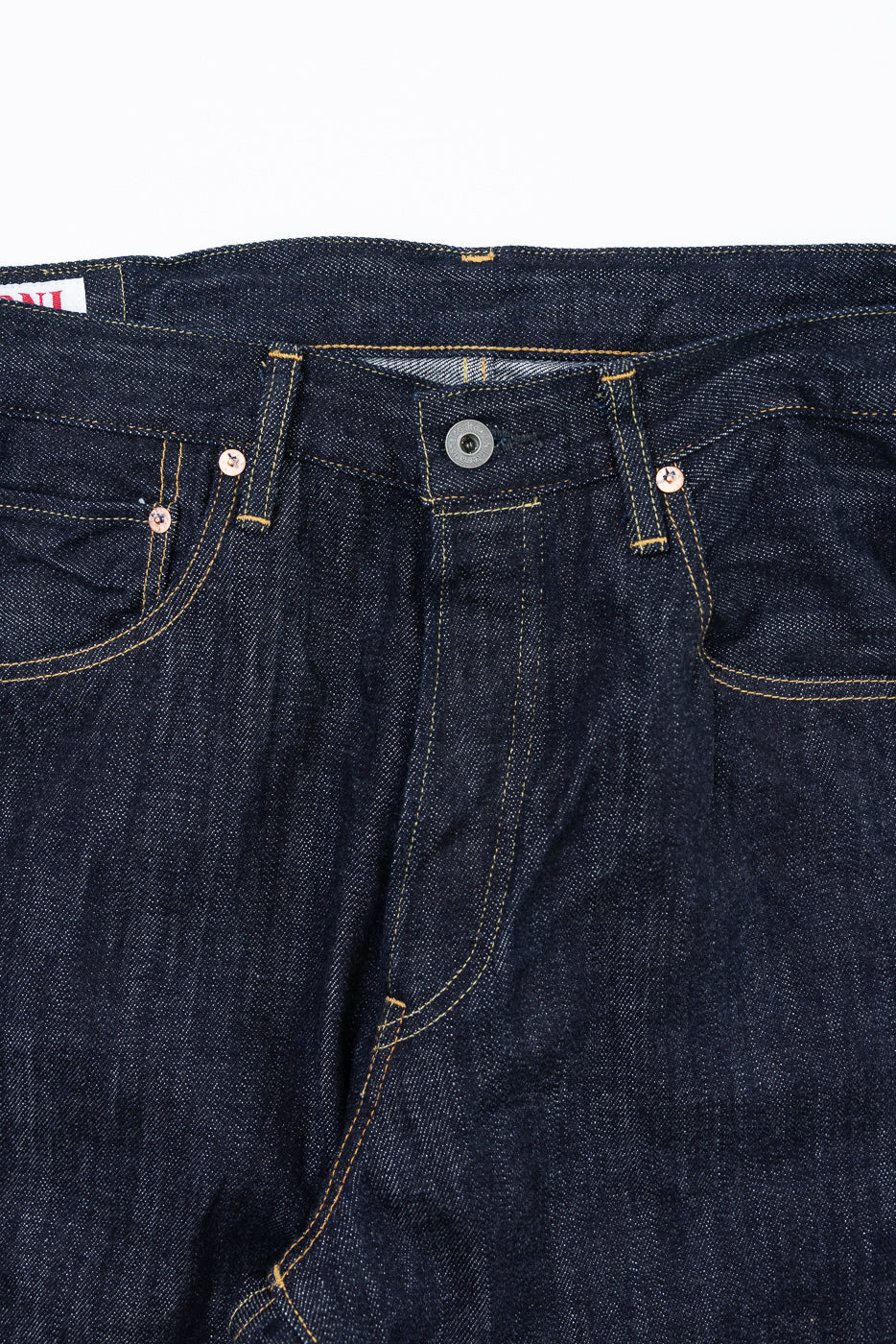 Close-up front view of dark selvedge denim jeans showing button fly, copper rivets, and yellow stitching detail