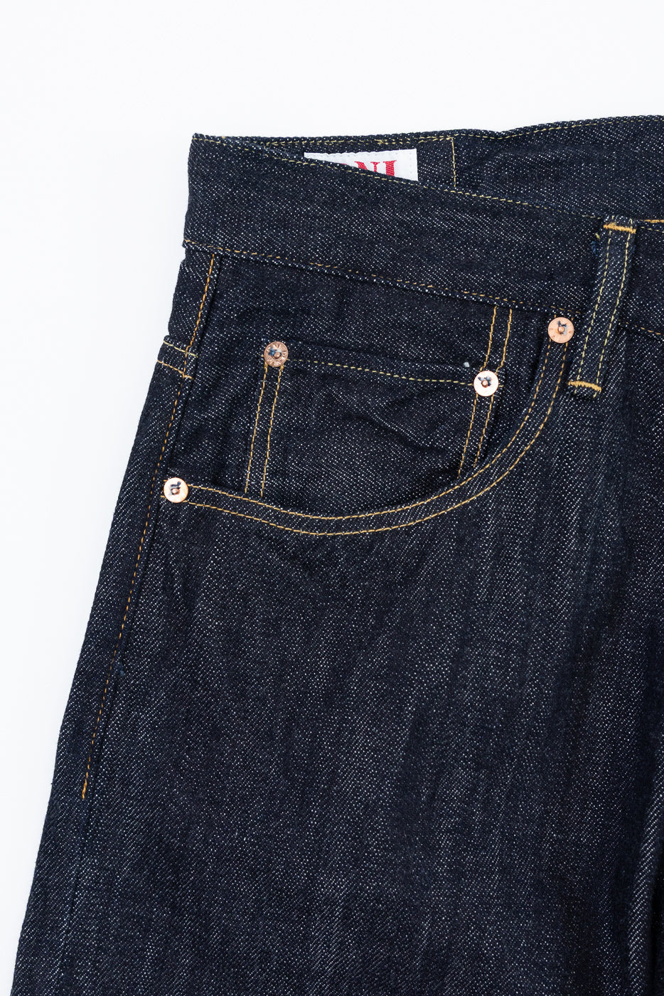 Close-up front view of dark selvedge denim jeans showing copper rivets, yellow stitching, and back pocket detail