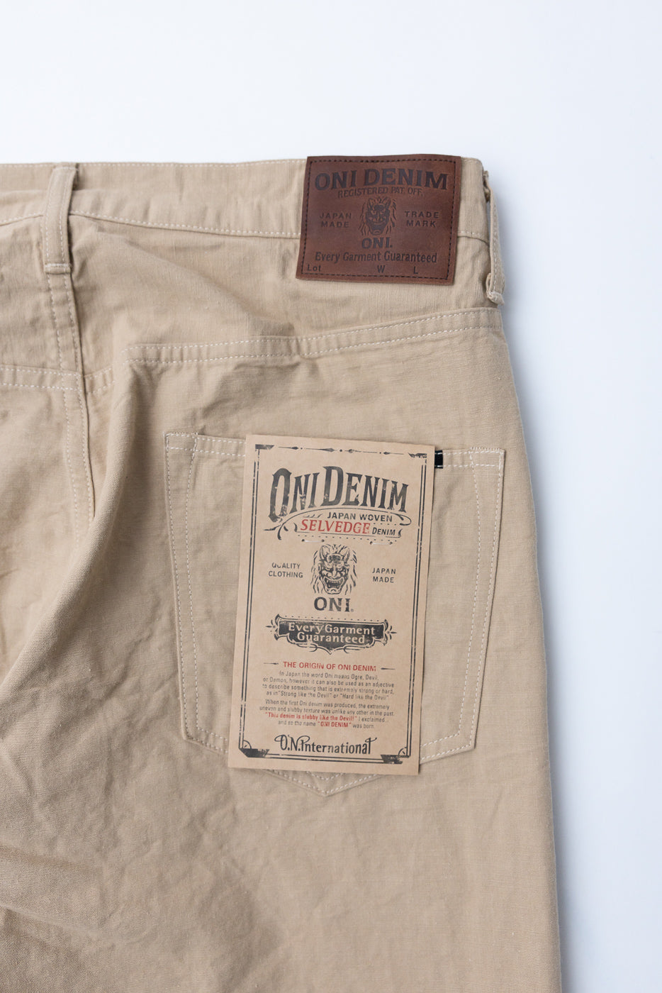 Close-up back view of beige selvedge denim pants showing ONI Denim leather waist tag and paper pocket label, Japan-made
