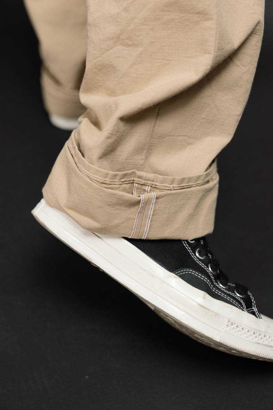 Close-up of male model's rolled khaki pant cuff with red/white stitching, paired with black sneakers, side view