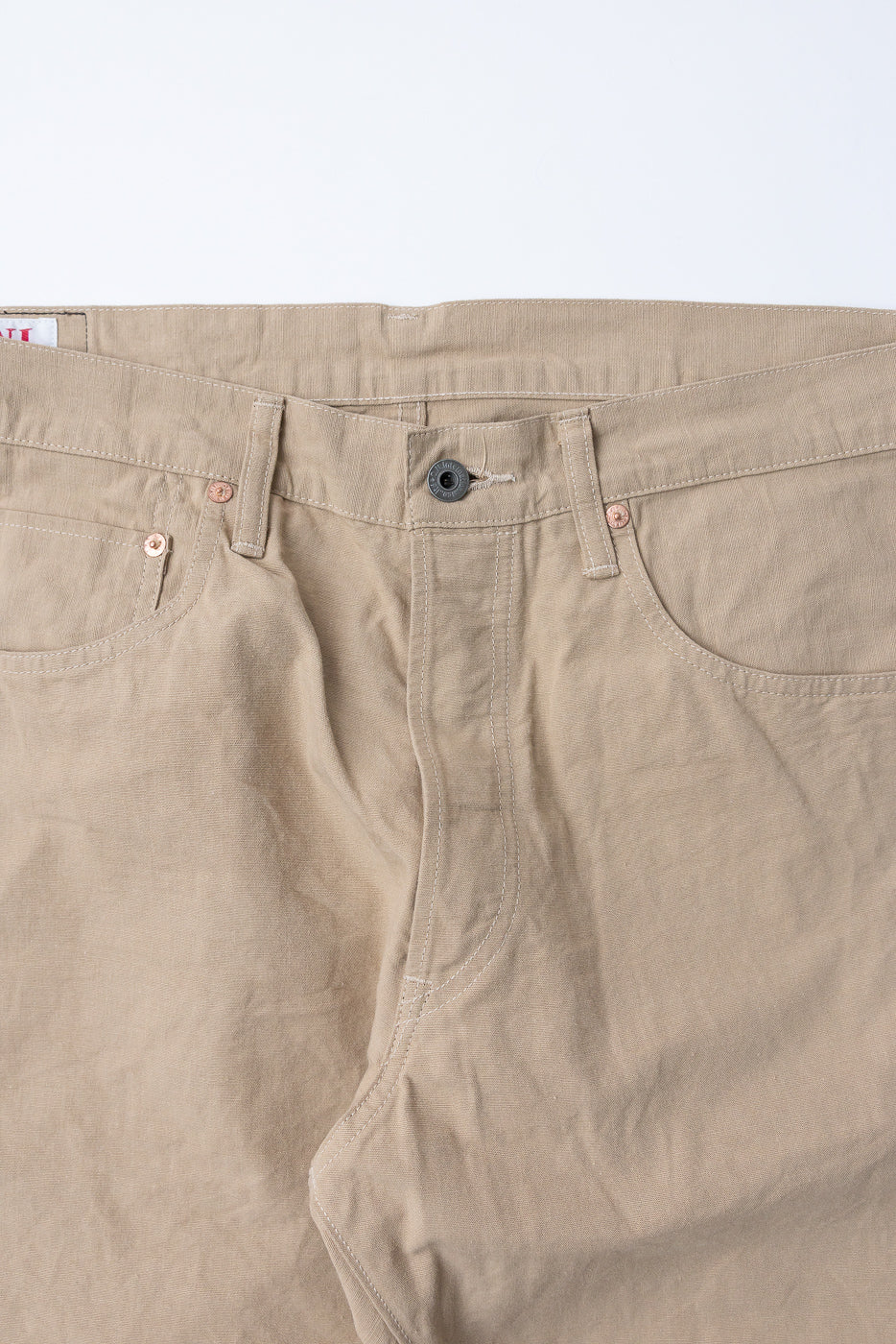 Close-up front view of beige casual pants showing button fly, copper rivets, and selvedge tag detail