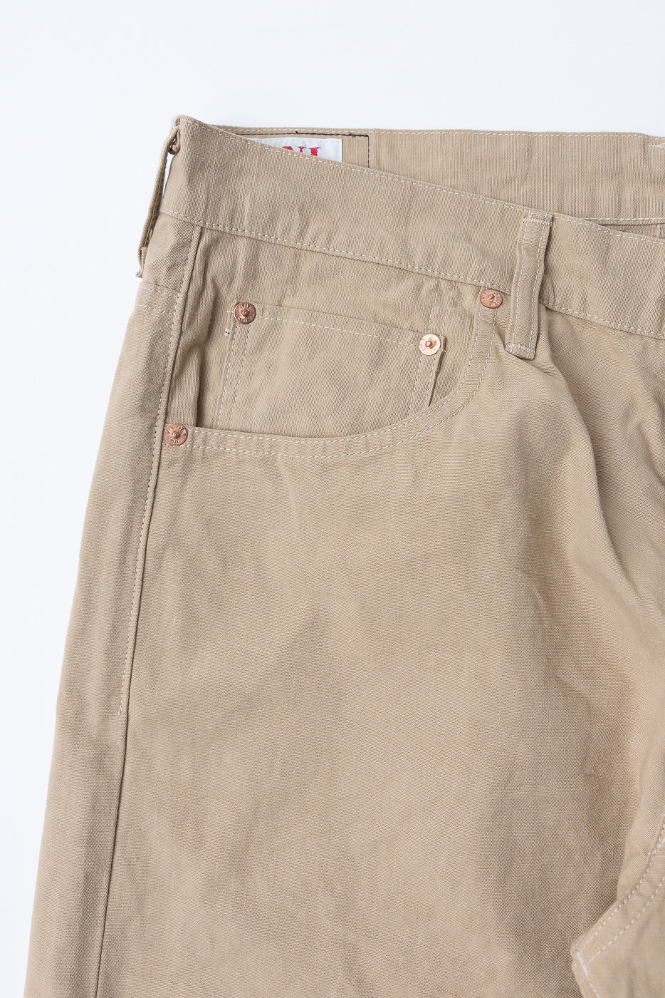 Close-up front view of beige casual pants showing front pocket, copper rivets, and waistband tag detail