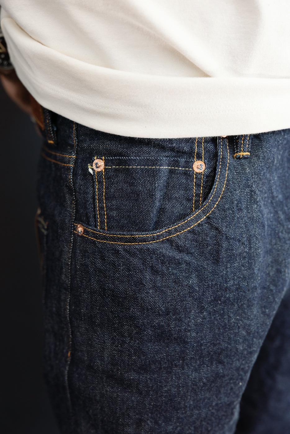 Close-up of the back right pocket of dark indigo selvedge denim jeans showing stitching and rivets.