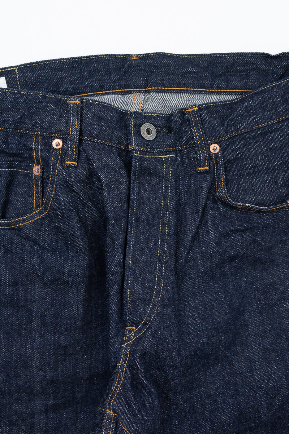 Close-up of the front waist and fly of dark indigo selvedge denim jeans with orange stitching.