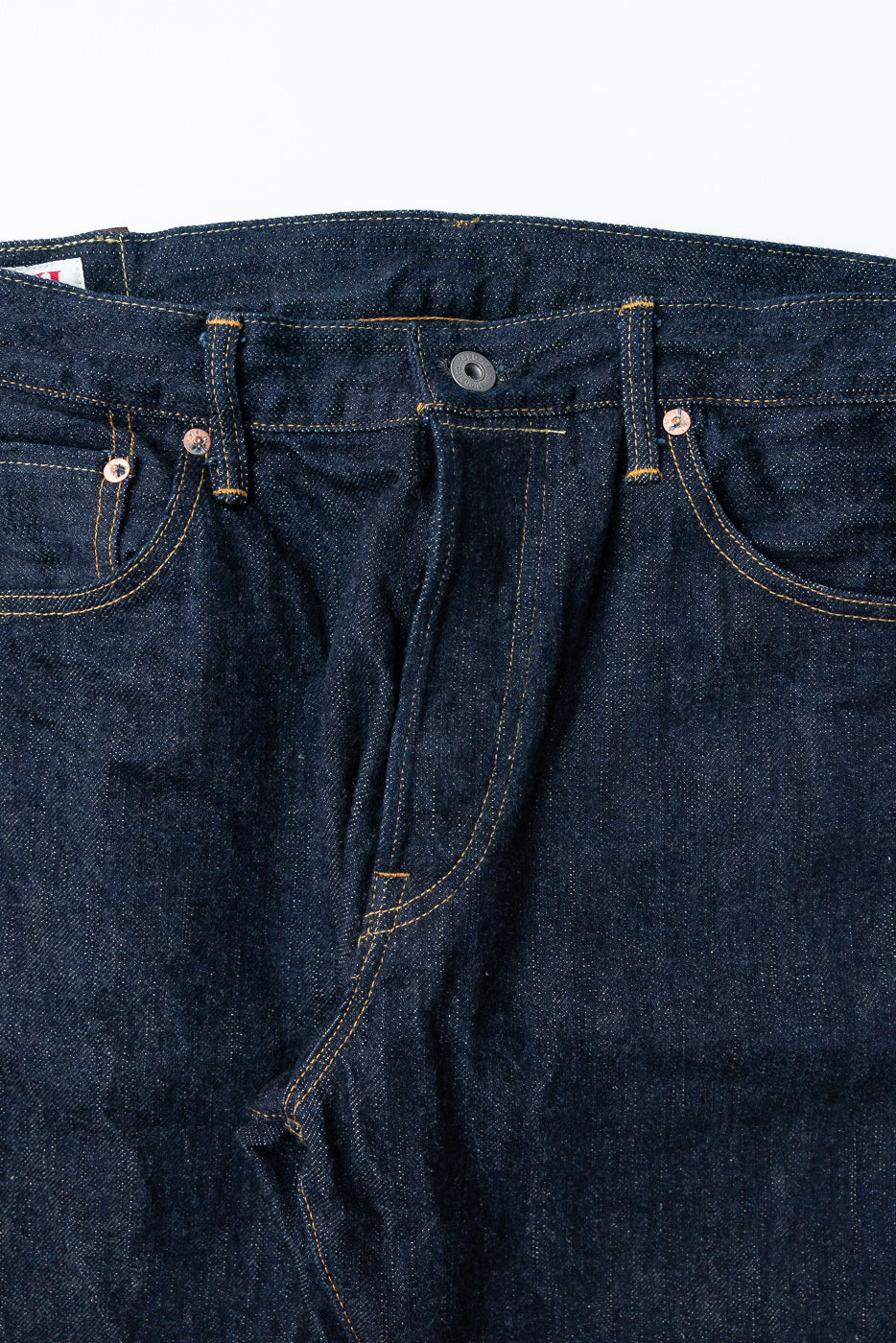 Close-up front view of raw selvedge denim jeans showing button fly, copper rivets, and yellow stitching detail.