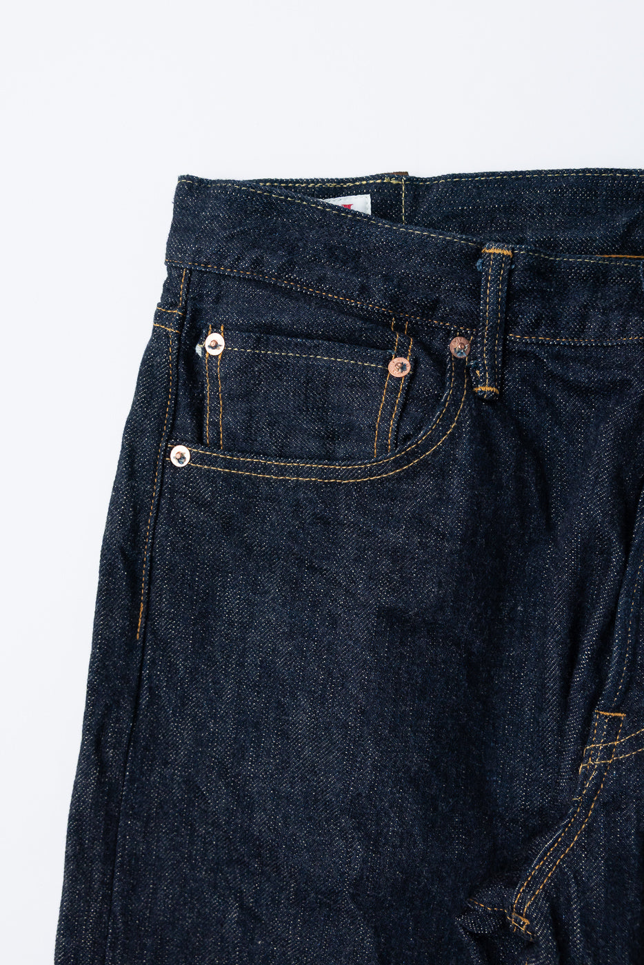 Close-up front view of dark selvedge denim jeans showing copper rivets, yellow stitching, and waistband tag detail