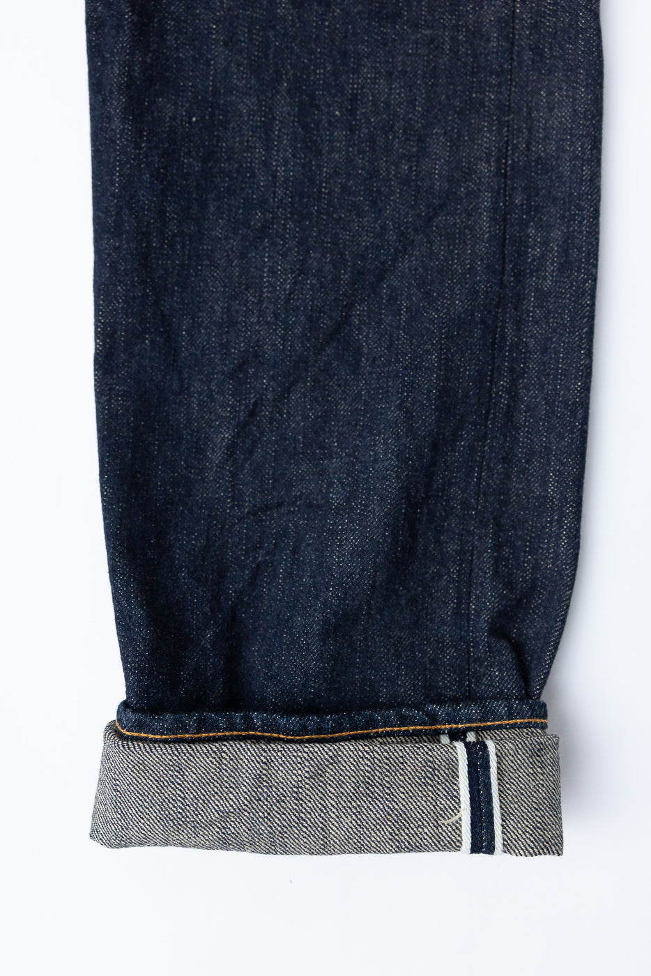 Close-up of dark selvedge denim jeans, front view showing cuffed hem with contrast stitching and fabric texture.