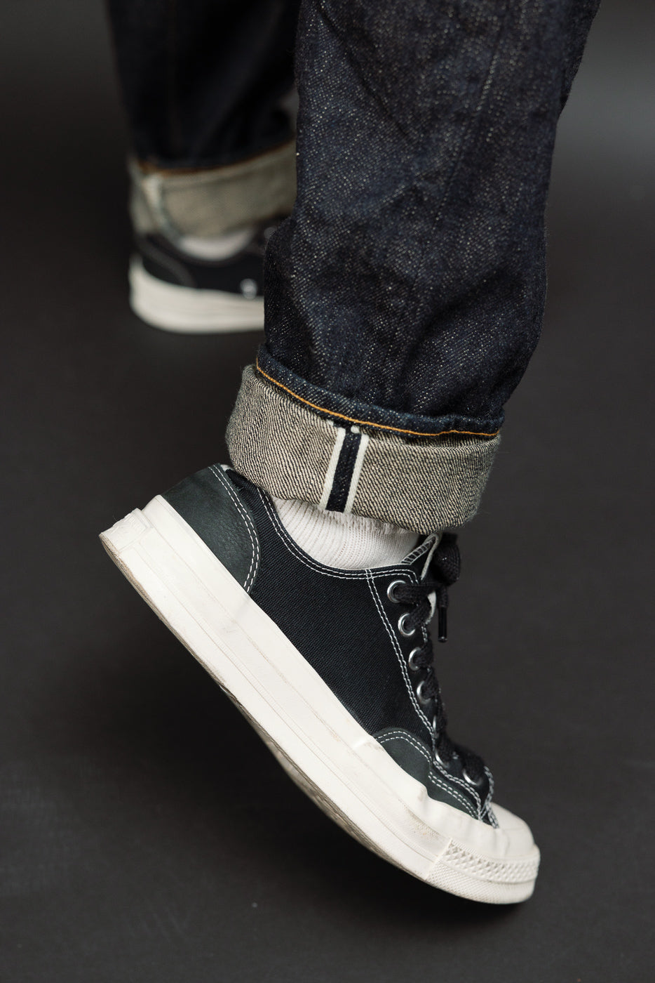 Close-up of male model's lower legs, showcasing dark selvedge denim with rolled cuffs and black sneakers.