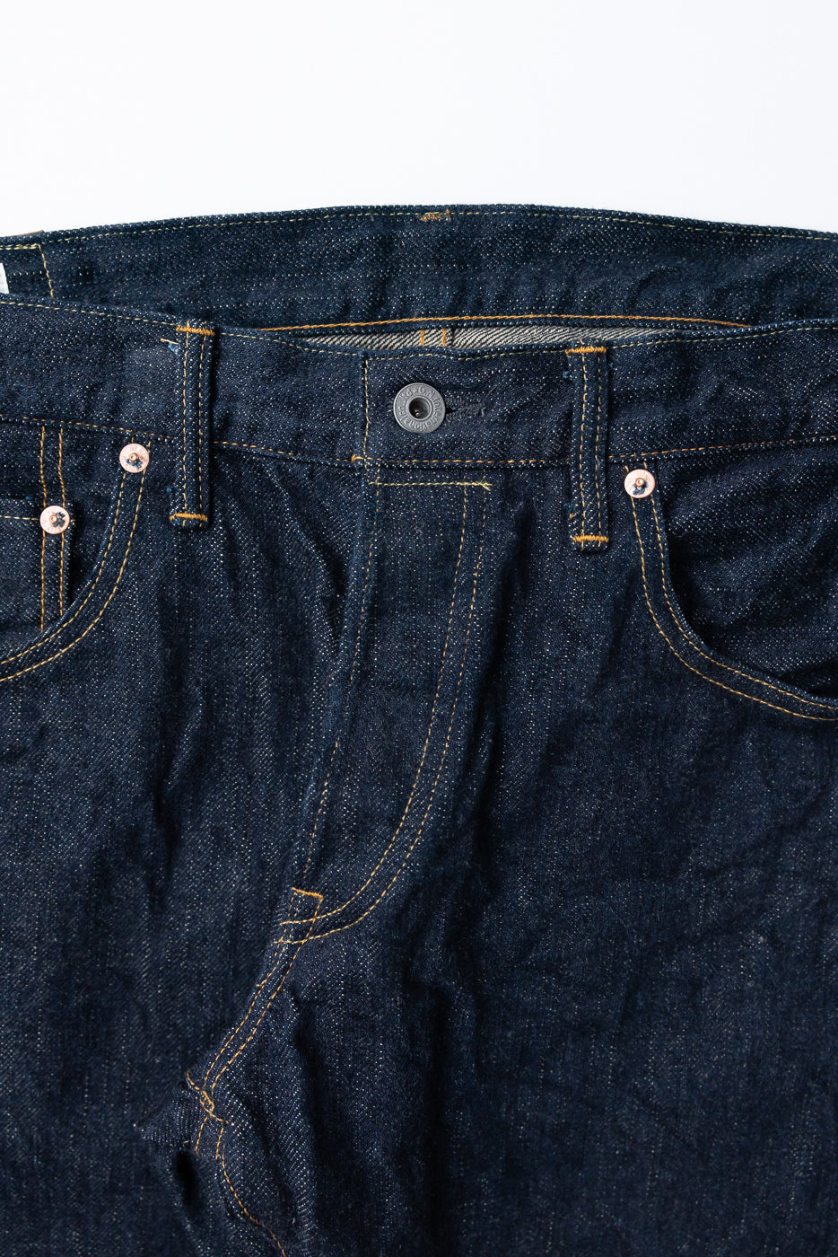 Close-up front view of raw selvedge denim jeans showing button fly, copper rivets, and contrast stitching
