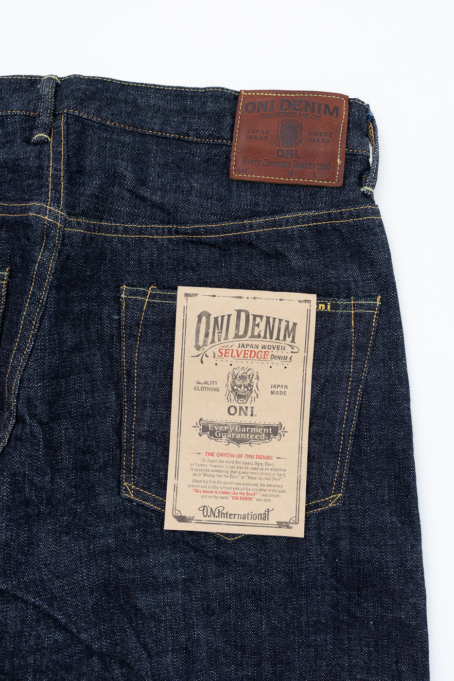 Close-up back view of Oni Denim selvedge jeans showing leather patch & paper tag, dark indigo fabric, yellow stitching
