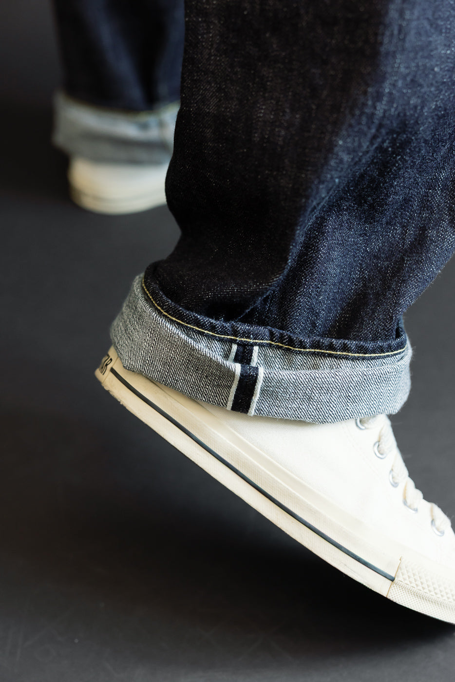 Close-up of male model's cuffed selvedge denim jeans, showing raw hem and white sneakers, front view