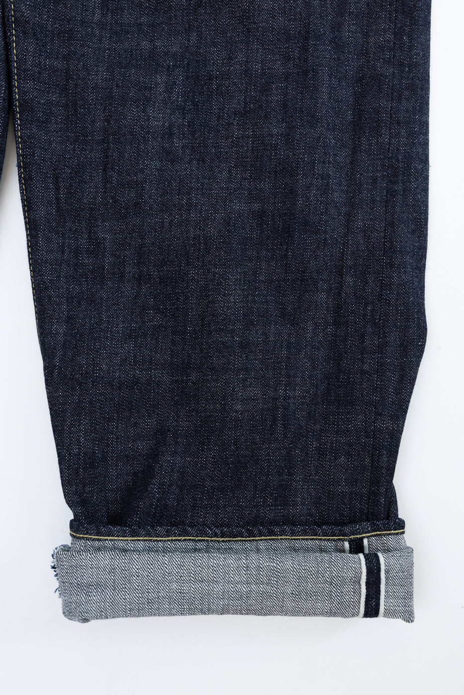 Close-up front view of dark selvedge denim jeans leg, showcasing raw hem, yellow stitching, and cuffed ankle detail