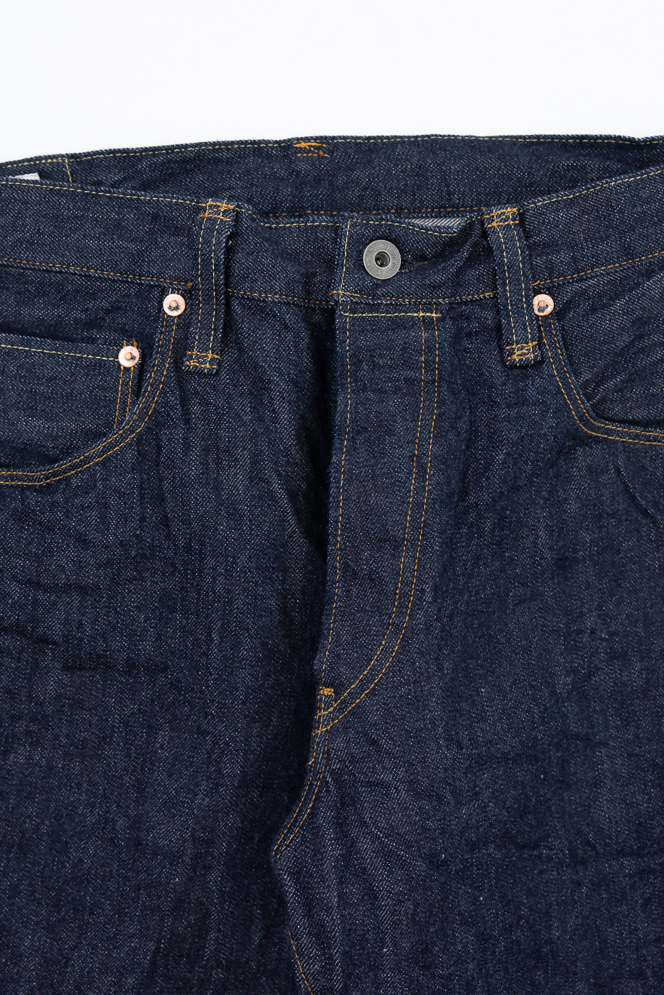 Close-up front view of dark indigo selvedge denim jeans showing the waistband, button, and stitching.