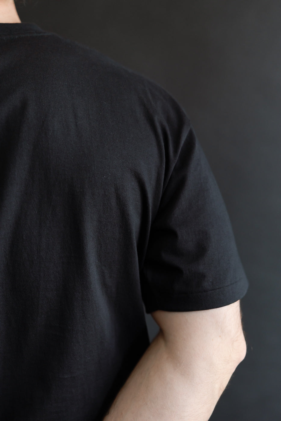 Close-up of back upper half of black t-shirt on male model, showing sleeve and fabric texture.