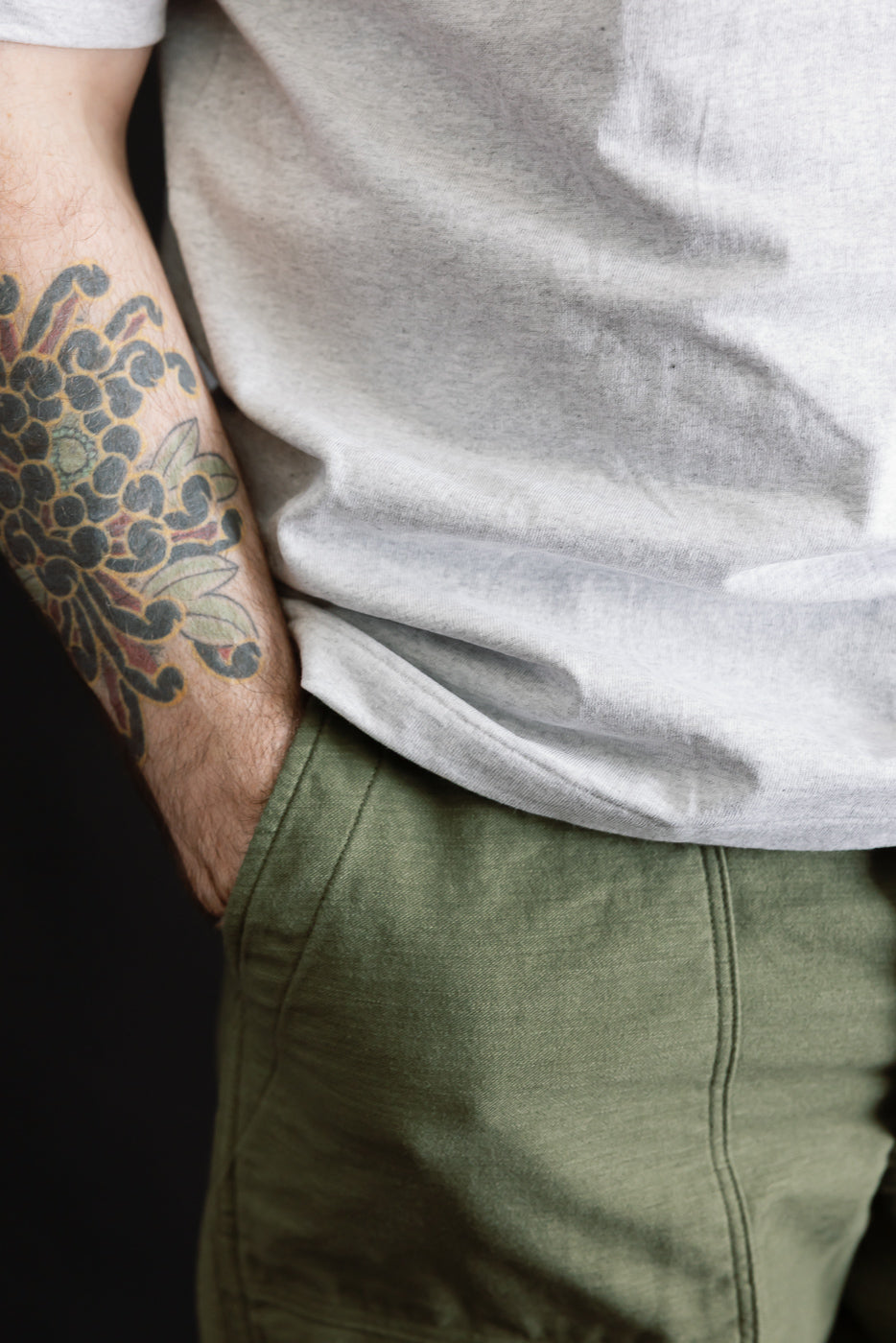Close-up of male model, 5'9, 165lbs, athletic build, wearing heather grey t-shirt front hem and olive pants, left side detail, tattooed arm visible.