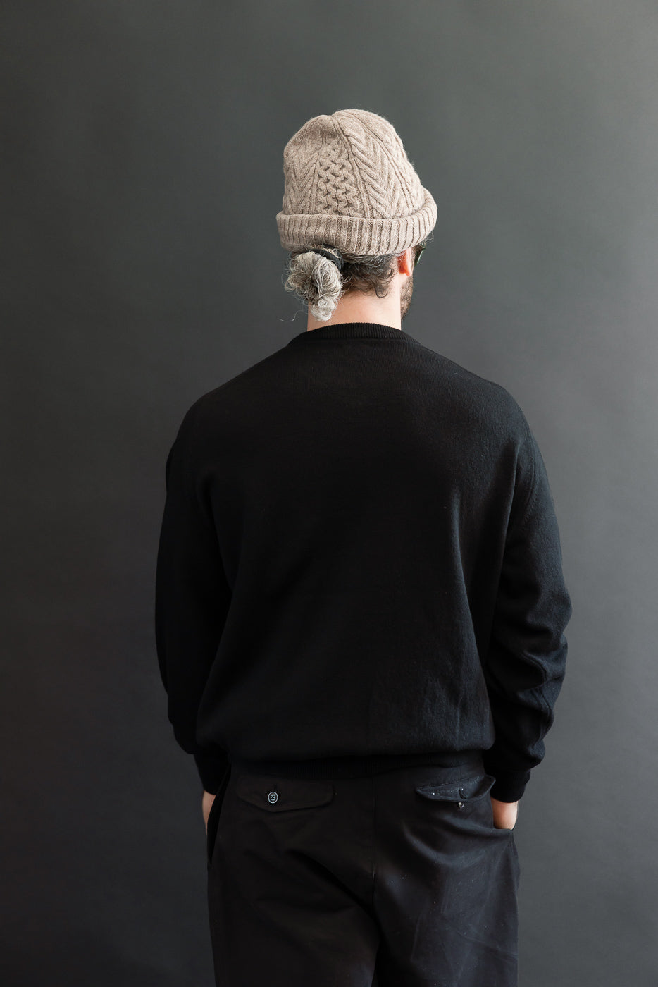 Back view of a male model wearing a black crewneck sweater and black trousers against a gray background.