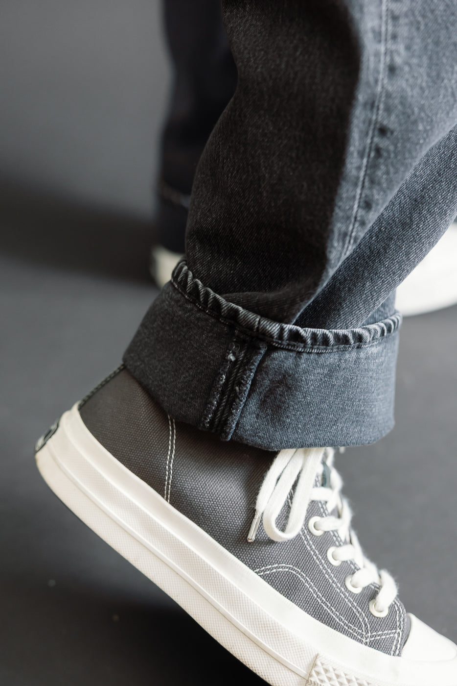 Close-up of the cuffed hem of dark wash denim jeans and grey canvas sneakers on a male model.