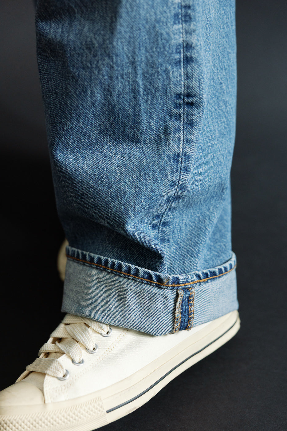 Close-up of male model's cuffed selvedge denim jeans, showing raw hem & contrast stitching with cream sneakers